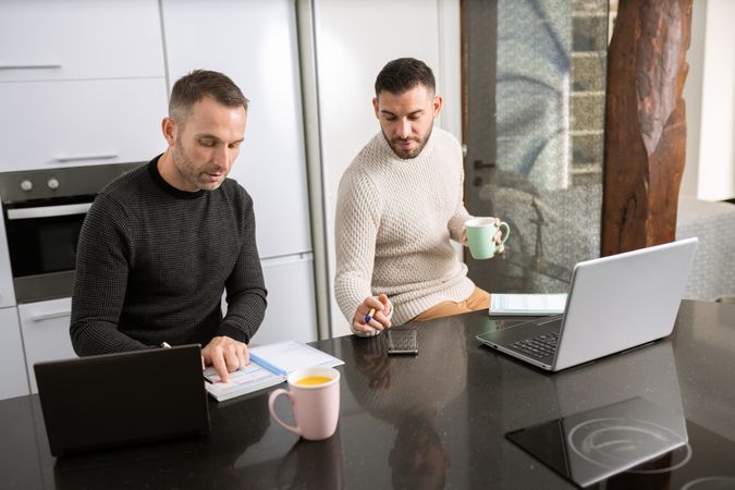 Two men with coffee working on laptops from their kitchen counter