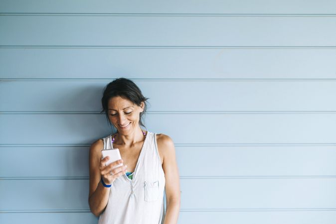 Smiling woman with ponytail checking phone while leaning on wood wall