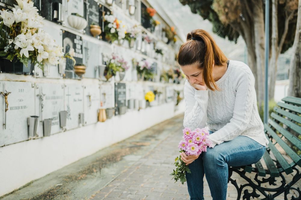 Crying woman witting on bench near grave wall in cemetery