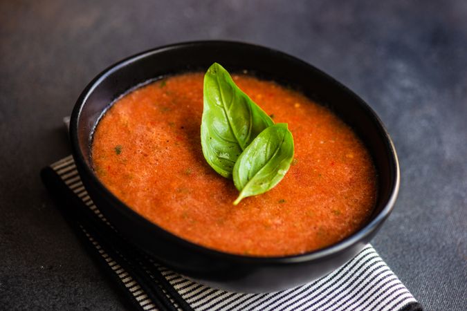 Dark bowl of gazpacho soup with basil leaves and wooden board