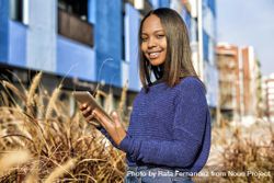 Happy female in sweater standing outside blue building in long grass with tablet 497pv0
