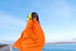 Trendy woman turning around in sunglasses, orange coat, yellow hoodie and jeans against the sea