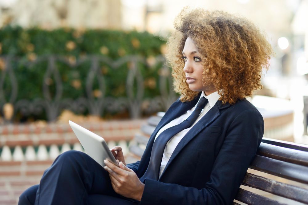 Woman in suit looking up from her digital tablet’s screen on outdoor bench