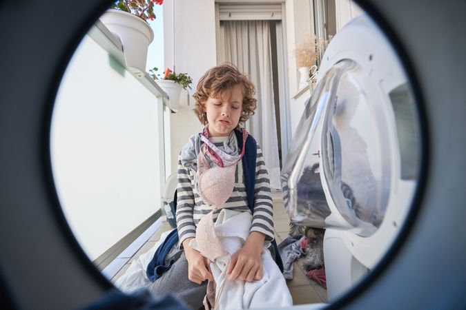 Upset boy with dirty clothes sitting near washing machine