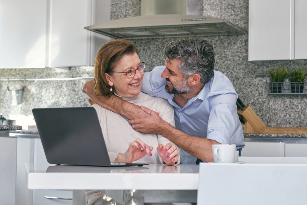 Adult son hugging senior mother at table with laptop