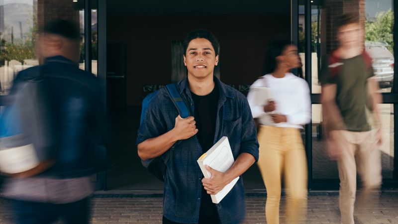 Confident student standing holding backpack and book looking at camera