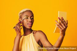 Black woman with short blonde hair holding a pocket mirror applying gold eyeshadow bD8aVb