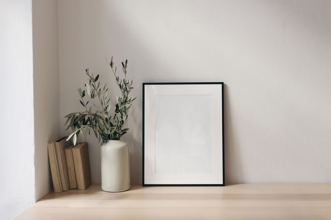 Elegant Mediterranean working space. Books, empty vertical black picture frame mockup. Wooden desk, table. Vase with olive branches. Home office concept. Scandinavian interior design.