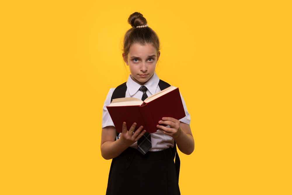 Serious schoolgirl holds open book while standing.