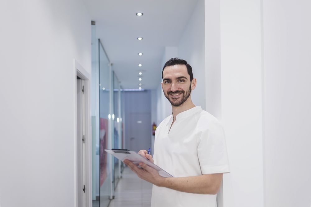 Portrait of a smiling doctor with beard holding a clipboard while leaning on wall in clinic corridor