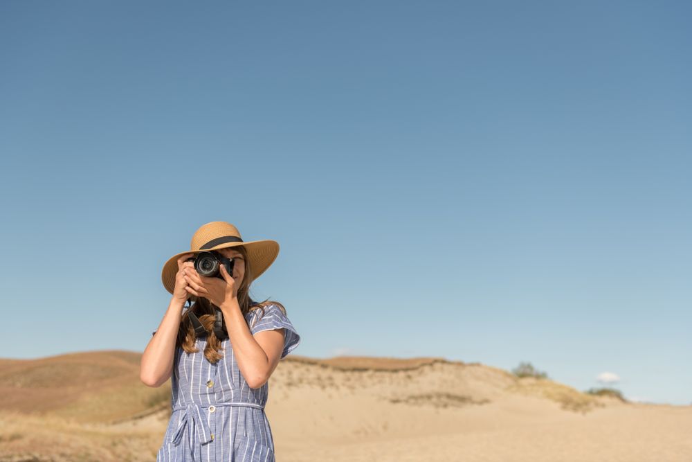 Back view of adult woman in straw hat and dress with camera taking photo on sand dune