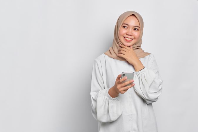 Asian female in headscarf smiling while thinking as she holds cell phone
