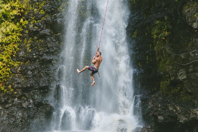 Man swinging on rope and jumping in water beside waterfall