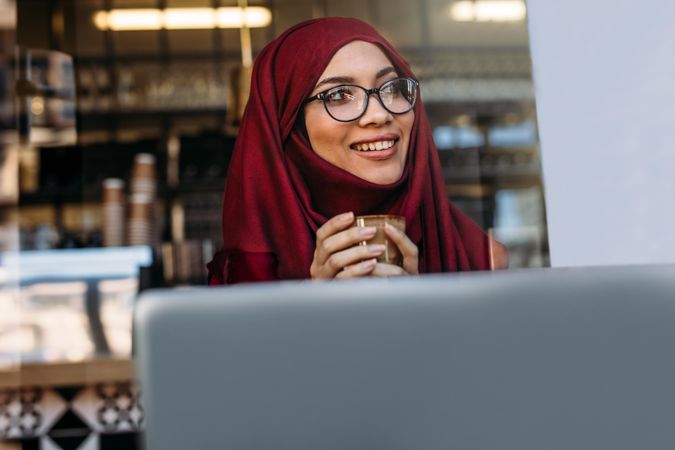 Woman in hijab and eyeglasses sitting at cafe with coffee in hand and looking away