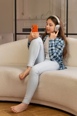 Girl sitting on sofa wearing headphones and using smartphone.