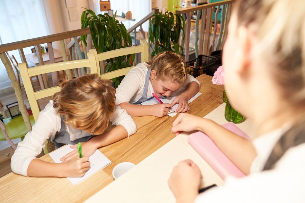 Girl in front of two children sitting in wooden chairs and with apron writing in the notebook