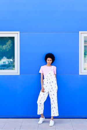 Woman in floral coveralls leaning against outside of blue building, copy space