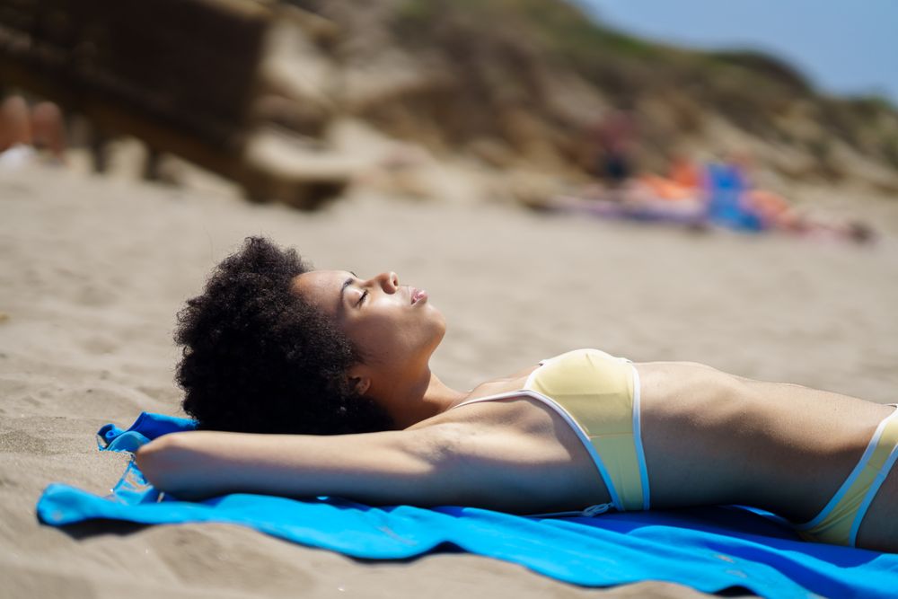 Female with curly hair in yellow bikini relaxing on blue towel on beach