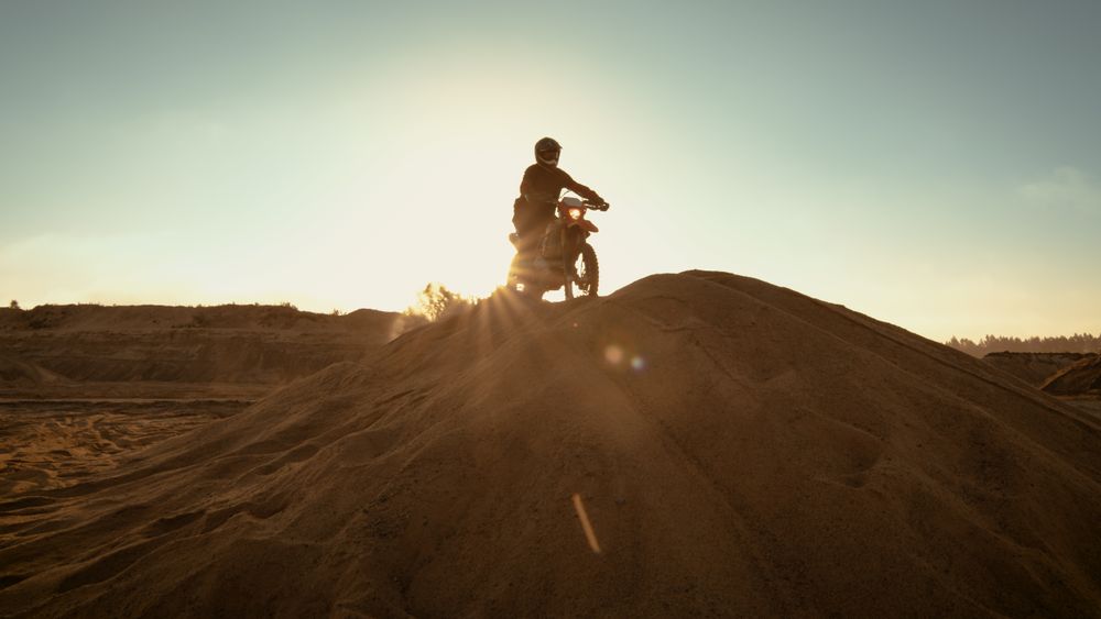 Professional Motocross Rider on FMX Motorcycle Drives on the Sand Dune and Stops There to Admire Scenic Sunsetting View.