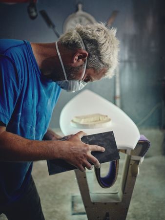 Serious surfboard shaper working using sandpaper in workshop