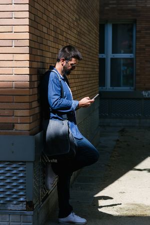 Man leaning on brick wall outside checking phone