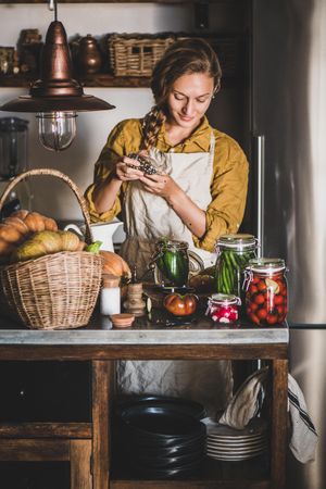 Young woman in rustic kitchen smiling putting peppercorn into jar of cucumbers for pickling