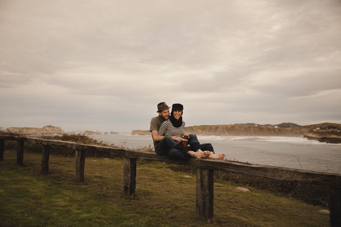 Young guy in hat near elegant woman playing on hand drum in cap on seat near coast of sea and cloudy sky
