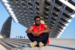 Black man wearing round sunglasses and red vest sitting cross legged outside