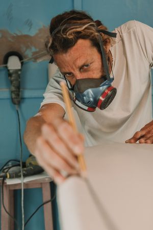 Man in respirator measuring surf board in workshop
