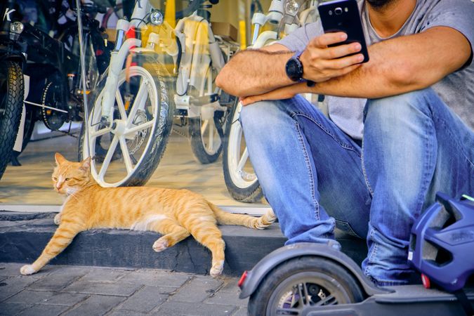 Man sitting on sidewalk using phone while cat lying beside