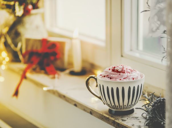 Mug with hot chocolate and whipped cream on window sill with candle, present and warm light