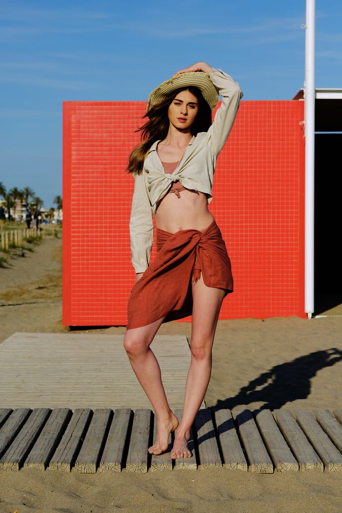 A long hair fashion model with a hat at the beach in front of a red wall