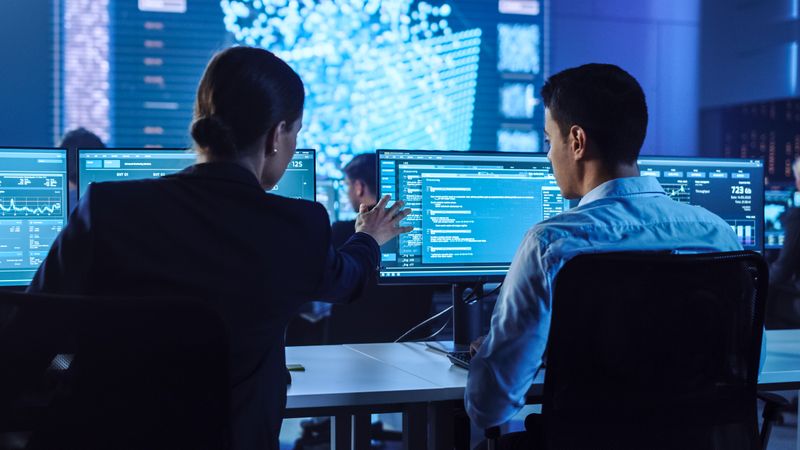 IT Specialists Collaborate in Dimly Lit Cybersecurity Operations Center. Multiple Screens Display Data, Charts, and Code. Blue-tinted Environment Emphasizes High-tech Atmosphere and Data Analysis.