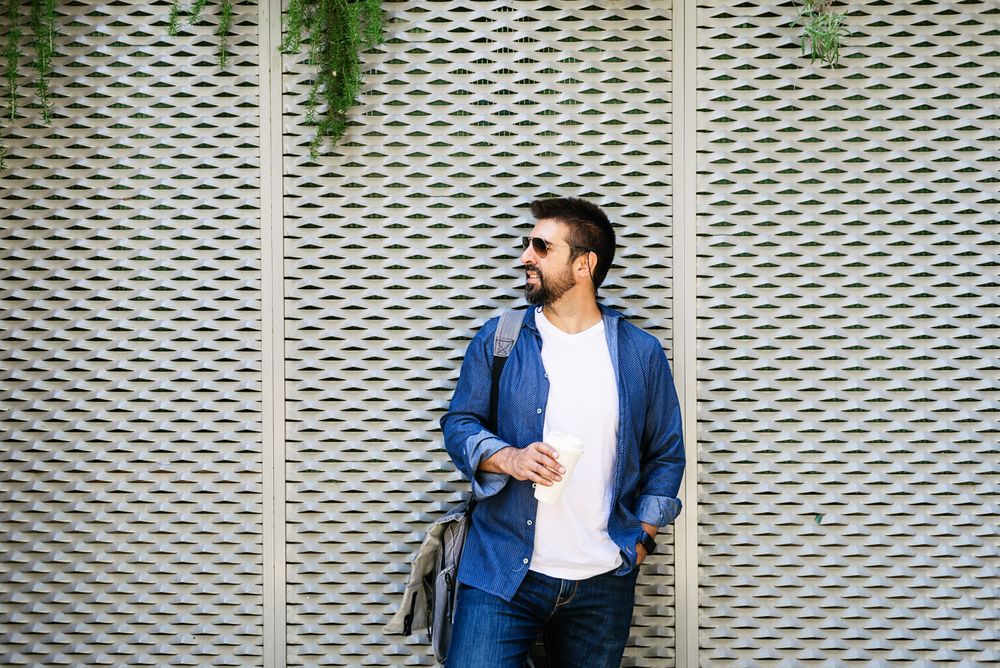 Man in denim leaning on fence outside with cup of coffee