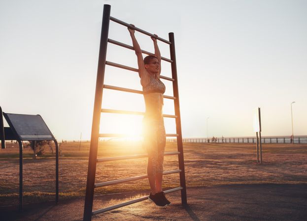 Full length shot of fit young woman in sportswear exercising on wall bars outdoors during sunset