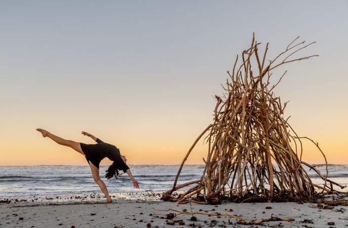 Young woman dancing on shore near water and wooden twigs placed for campfire