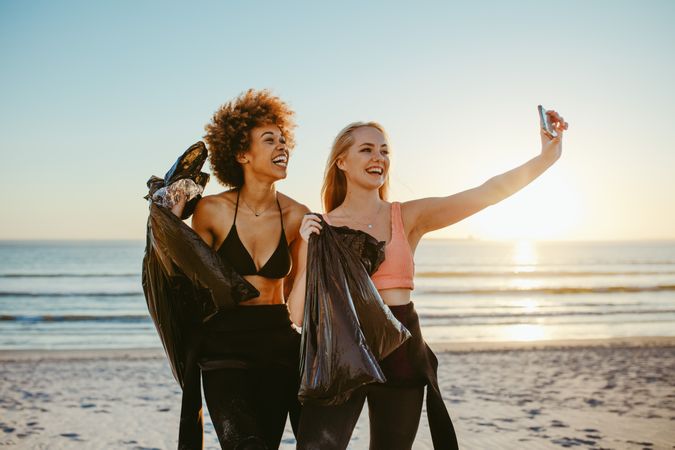 Two young women holding garbage bags and taking selfie after cleaning the beach area