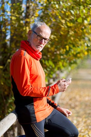 Older male checking phone sitting on wooden fence in park