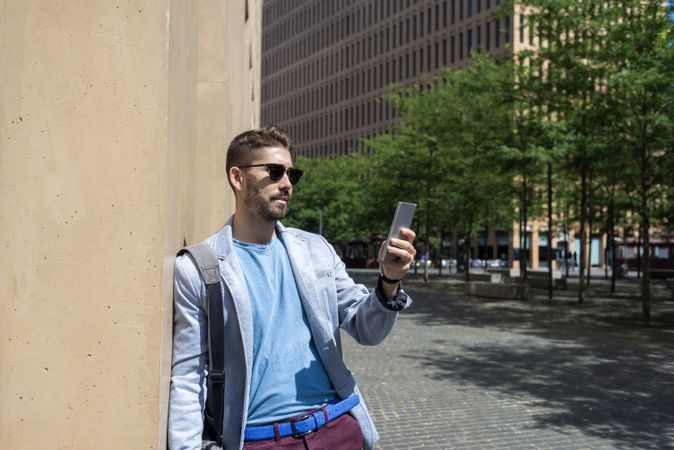 Bearded man in blazer leaning on a building wall outside taking a video call