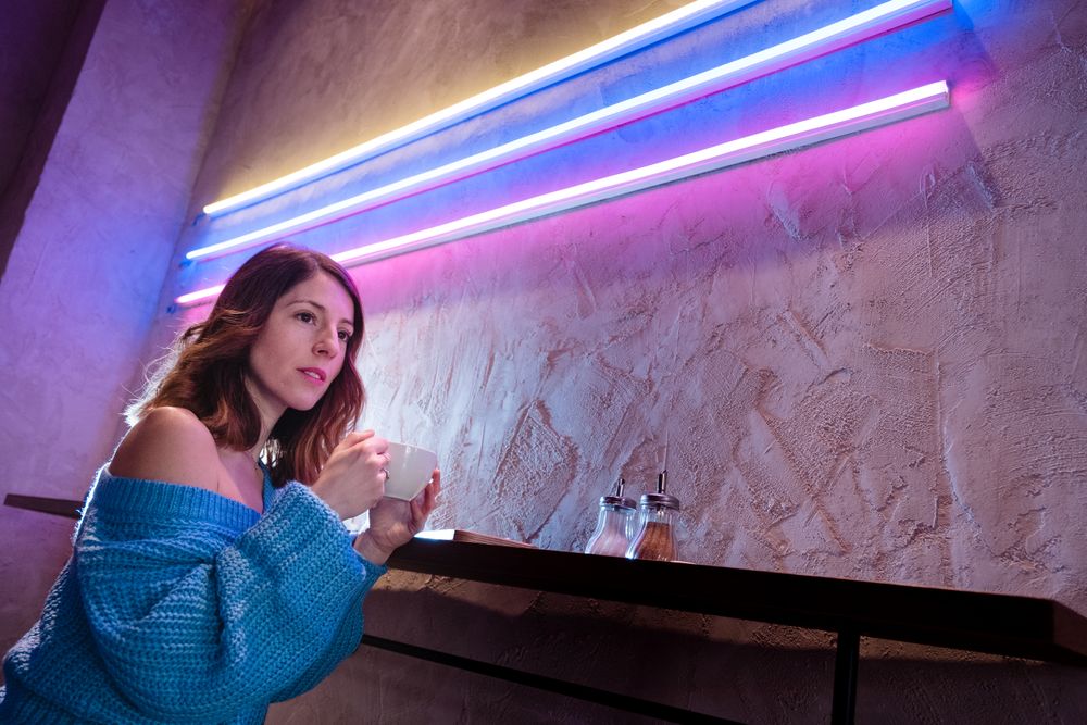 Positive woman with cup at table near wall with neon lamps