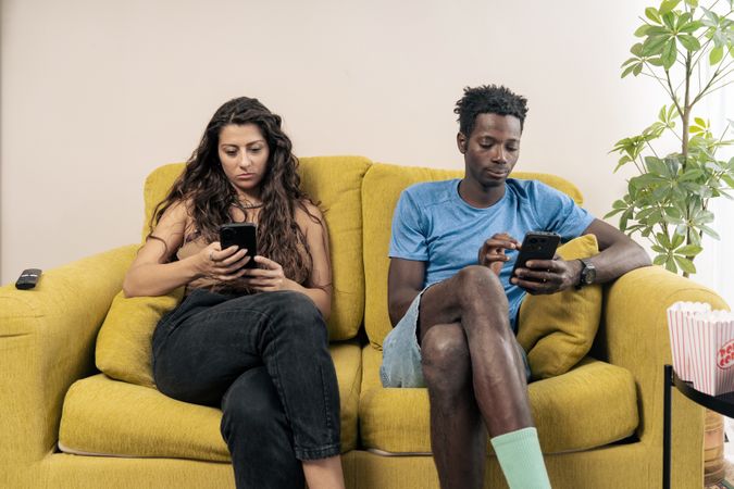 A young couple sits far apart on a couch, each staring silently at their own mobile phone, illustrating digital isolation.