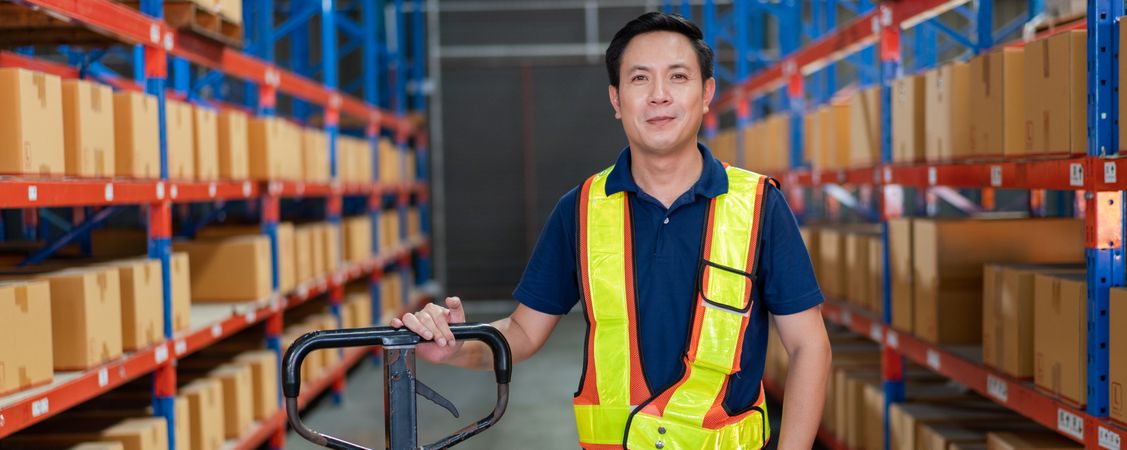 Smiling Asian warehouse worker with trolley in safety gear