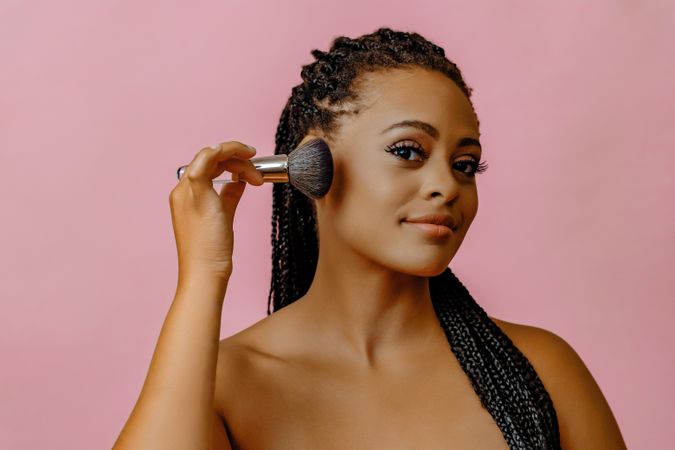 Black woman looking at camera while applying product with make up brush to her cheek