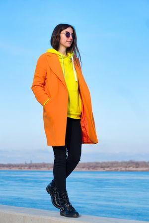 Woman in sunglasses, orange coat, yellow hoodie and jeans against the beach background