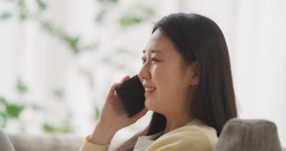 Sitting on the Sofa in Her Cozy Apartment, Smiling South Korean Woman is Having a Thoughtful Phone Conversation