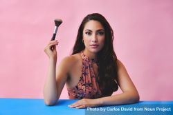 Hispanic woman with long brown hair holding large make up brush up and looking at camera 48yEZb