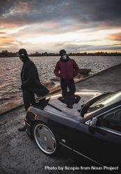 Two men with masks standing beside dark car by shoreline at sunset 0gMNM4