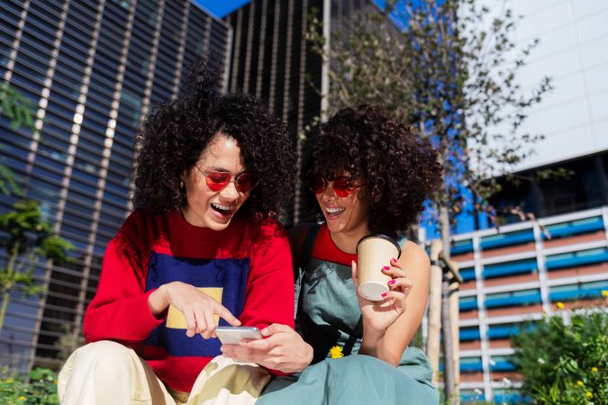 Two female friends sitting on bench outside checking phone together with coffee