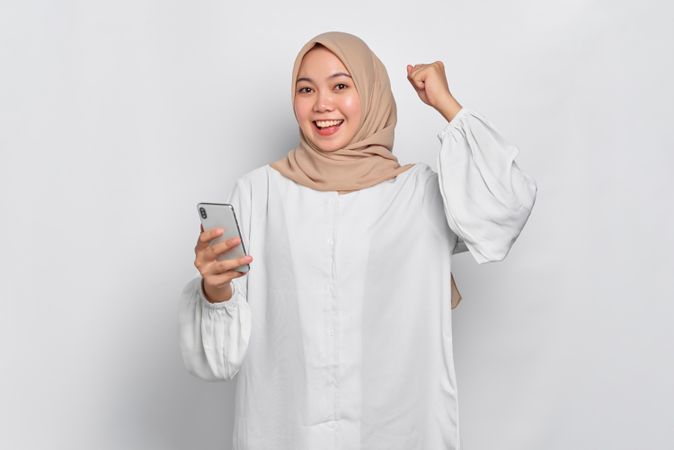 Asian female in headscarf smiling while celebrating good news as she holds cell phone
