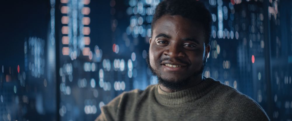 Close Up Portrait of Black Businessman Posing Next to Window in Big City Office with Skyscrapers Late At Night.